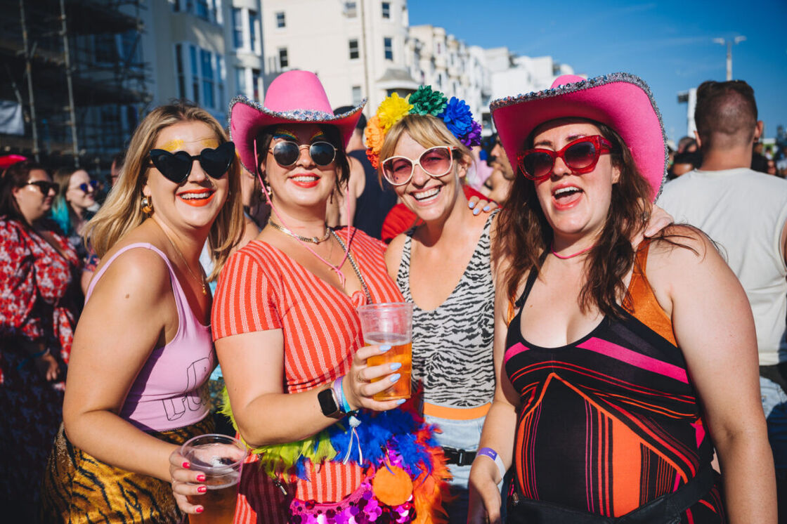 Four female presenting people wearing colorful hats and outfits smile at the camera