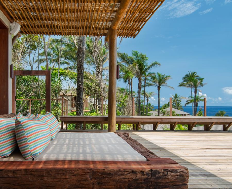 Bright blue sky with the ocean in the background. In the foreground a bed out on a large tropical patio. Photo via The Barracuda