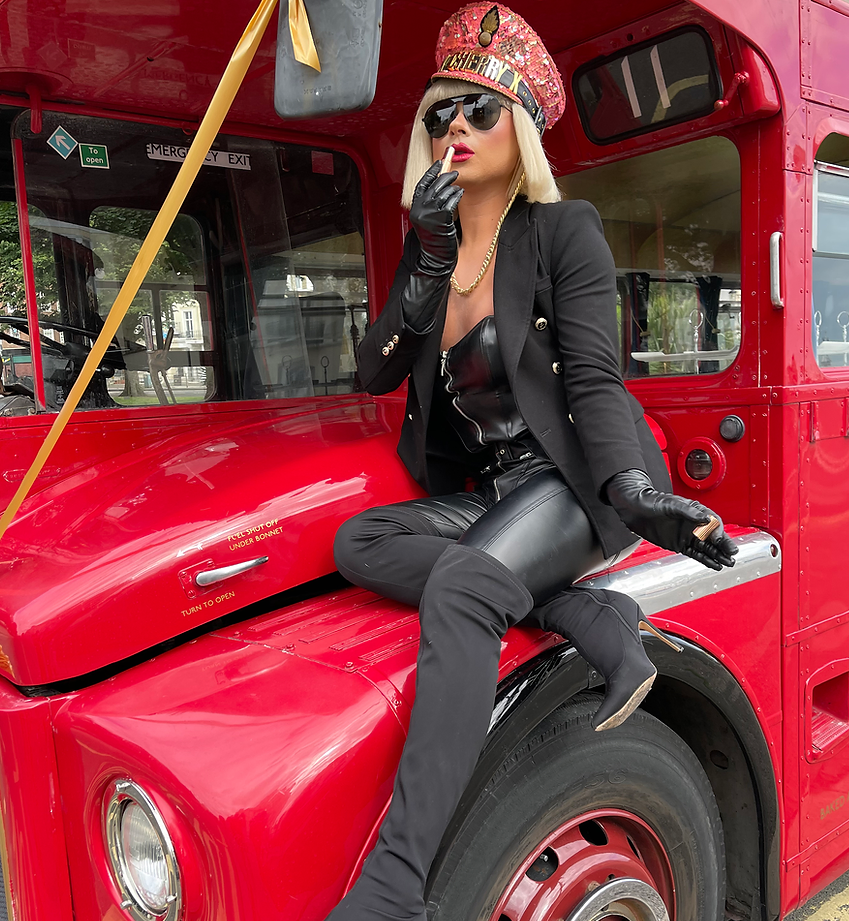 Gorgeous drag queen in a black suit and red, bedazzled hat applies lipstick while sitting on the hood of a red bus