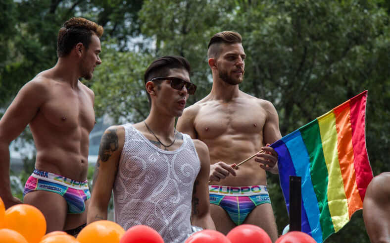 Two shirtless Mexican men in speedos and a man in sunglasses and tank between them on a float. 