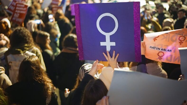 The backs of female marchers and one is holding a purple sign of the female symbol. 