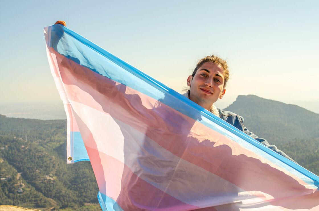 a transgender person holding a transgender pride flag on a hike in the mountains
