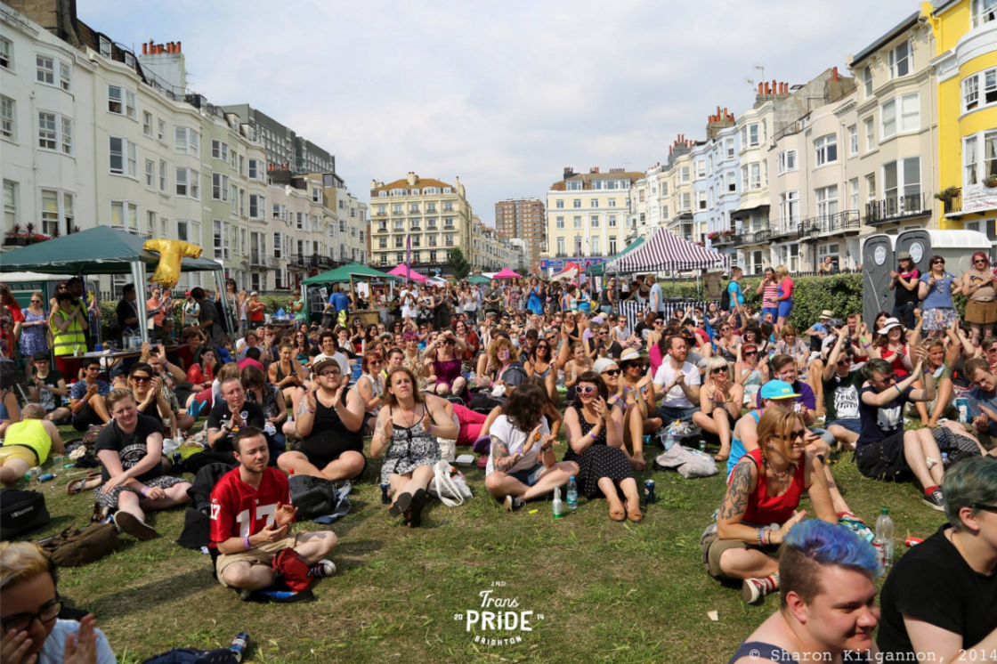 A large crowd sits on a lawn surrounded by buildings