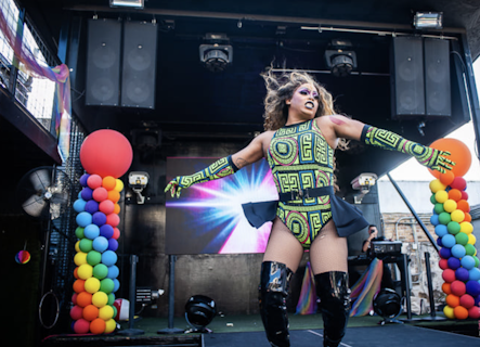 A fierce drag queen performs on stage in front of rainbow balloons