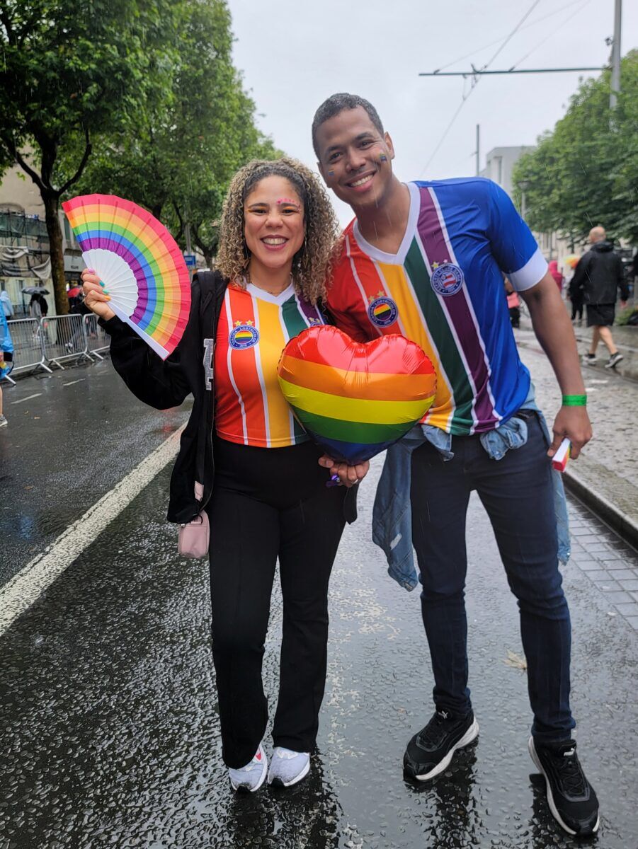 Two people in matching rainbow shirts stand on the rainy street