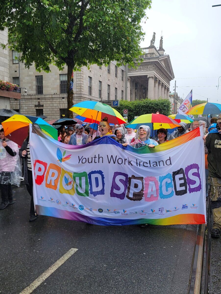 A group holding 'Proud Spaces' sign walks down the street