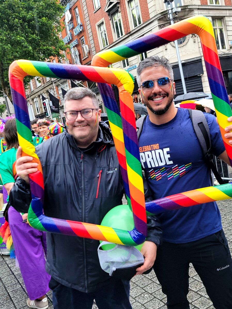 Two men stand with blow up rainbow picture frames around their faces