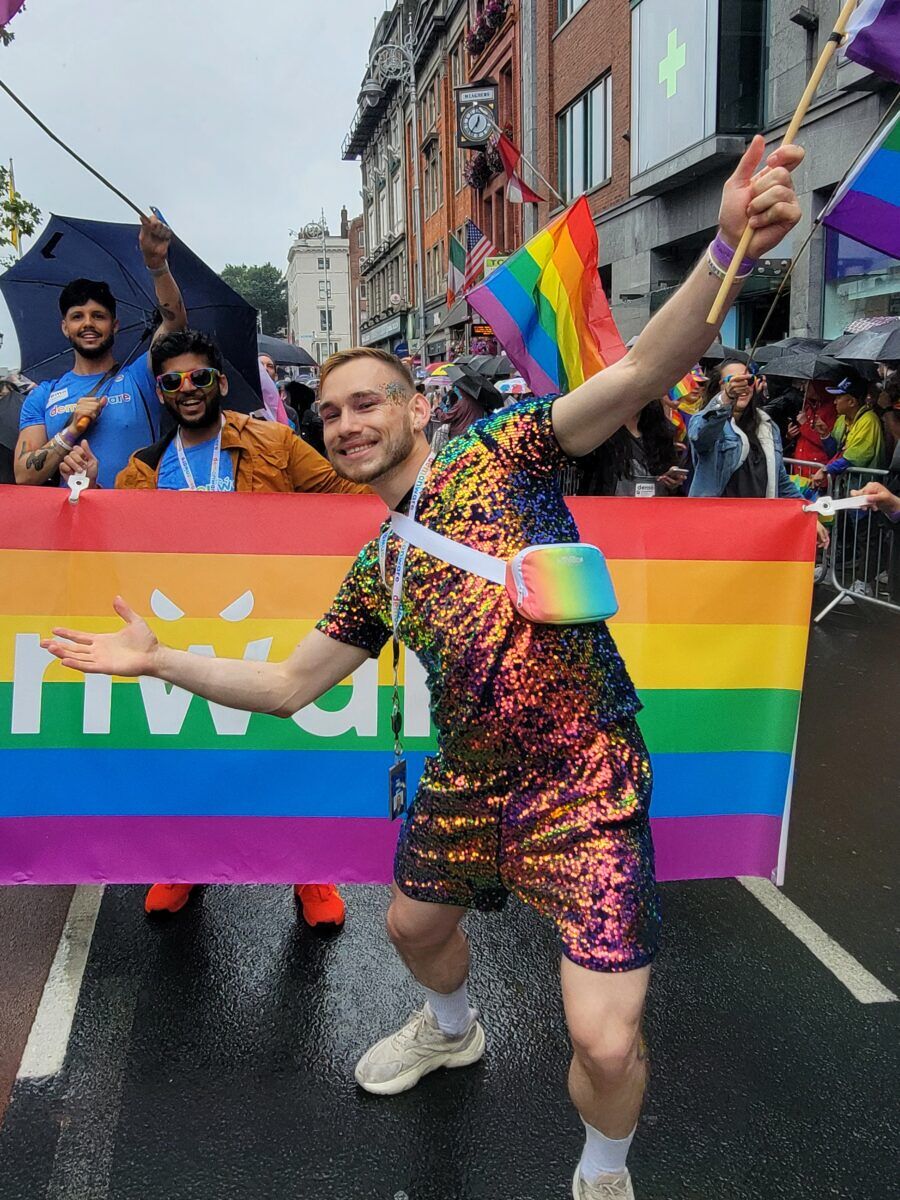 A man in a sequined shirt and matching shorts stands in front of a rainbow sign on the street