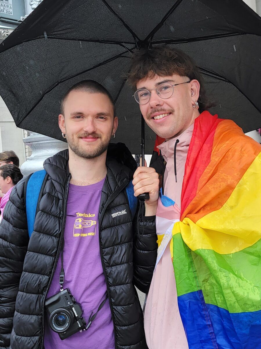 Two men stand under a black umbrella