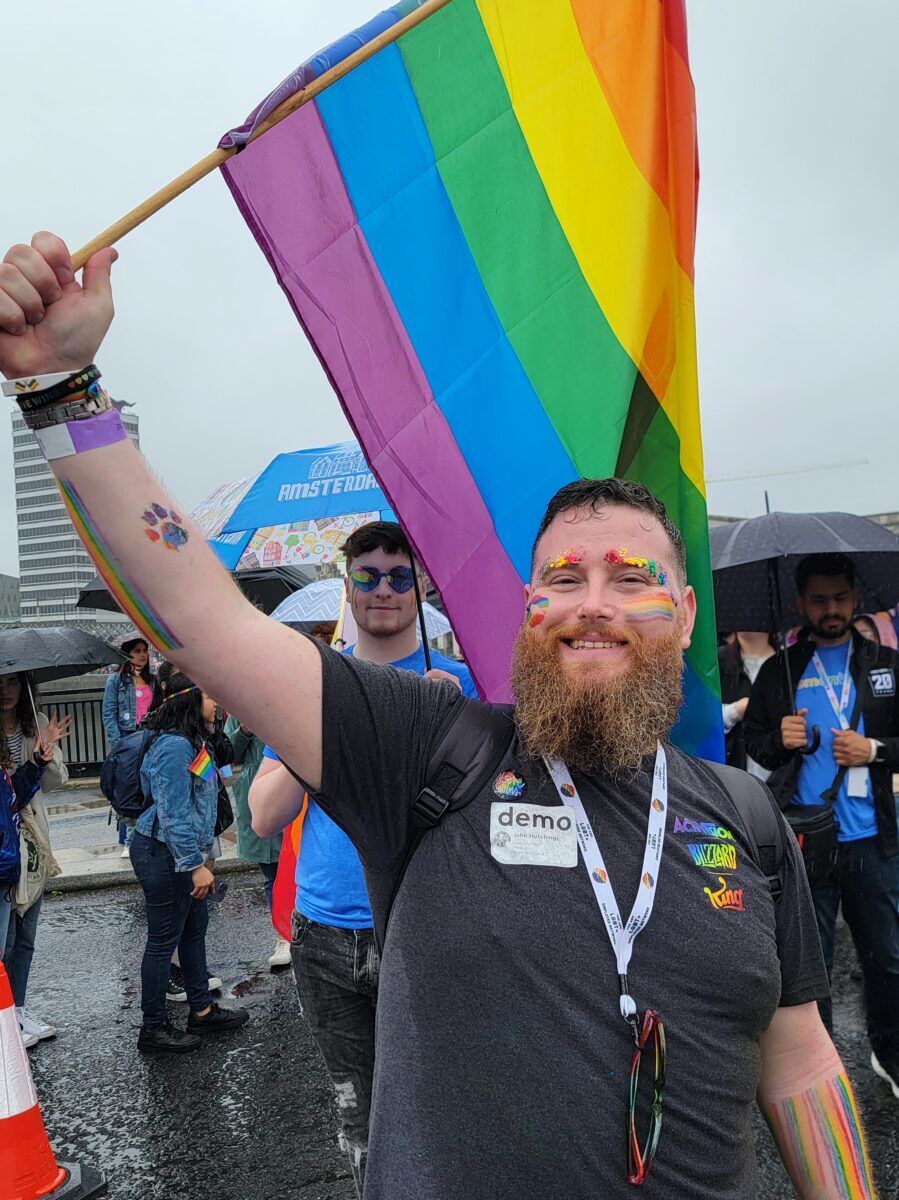 A bearded man stands smiling with a rainbow flag