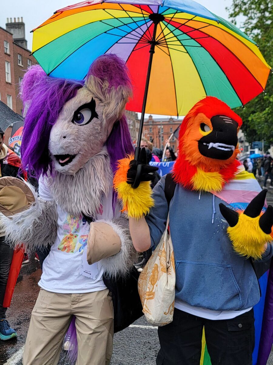 Two people in furry costumes stand under a rainbow umbrella