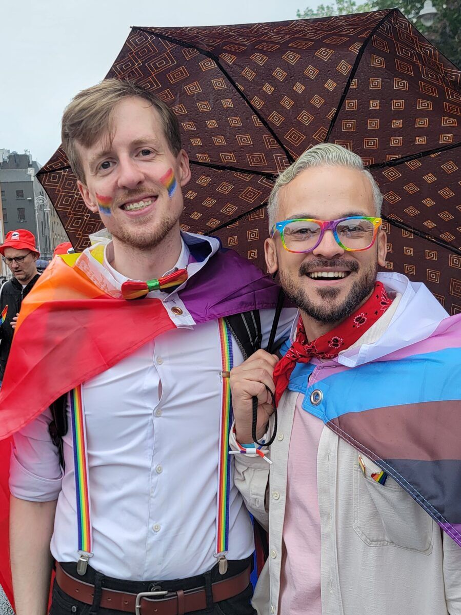 Two men smile at the camera while under an umbrella