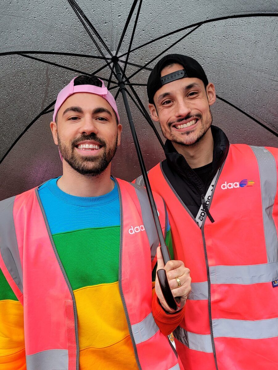 Two men with backwards baseball caps stand under an umbrella