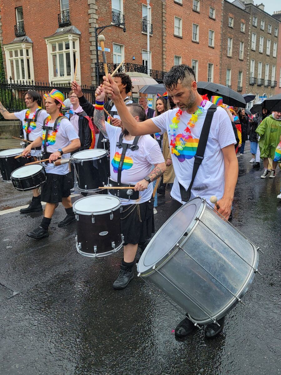 A rain-soaked drumline with rainbow attire leads a group down the street