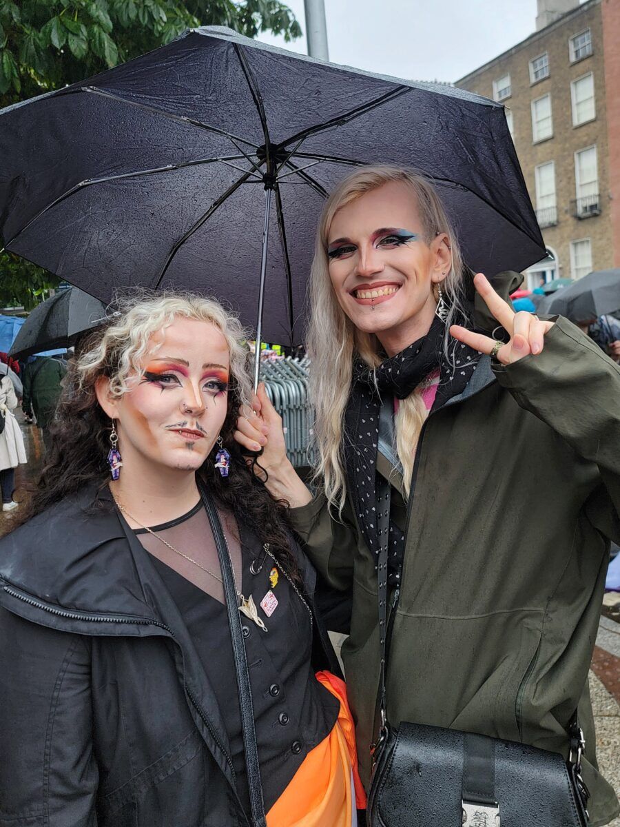 Two people with bright makeup stand under an umbrella