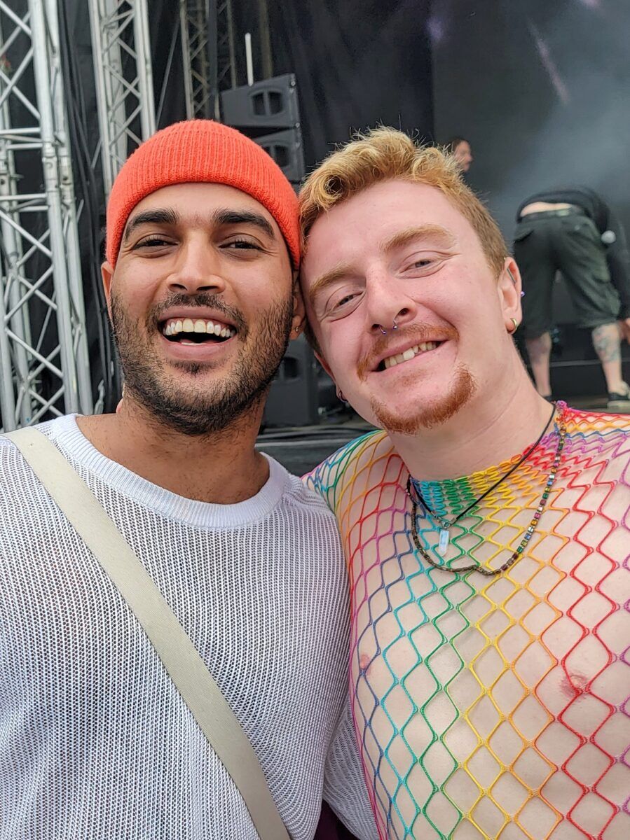 Two men, one in a mesh rainbow top smile at the camera