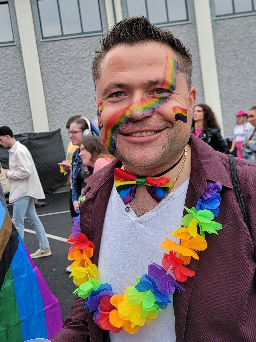 A man with a rainbow painted on his face and rainbow bowtie smiles at the camera