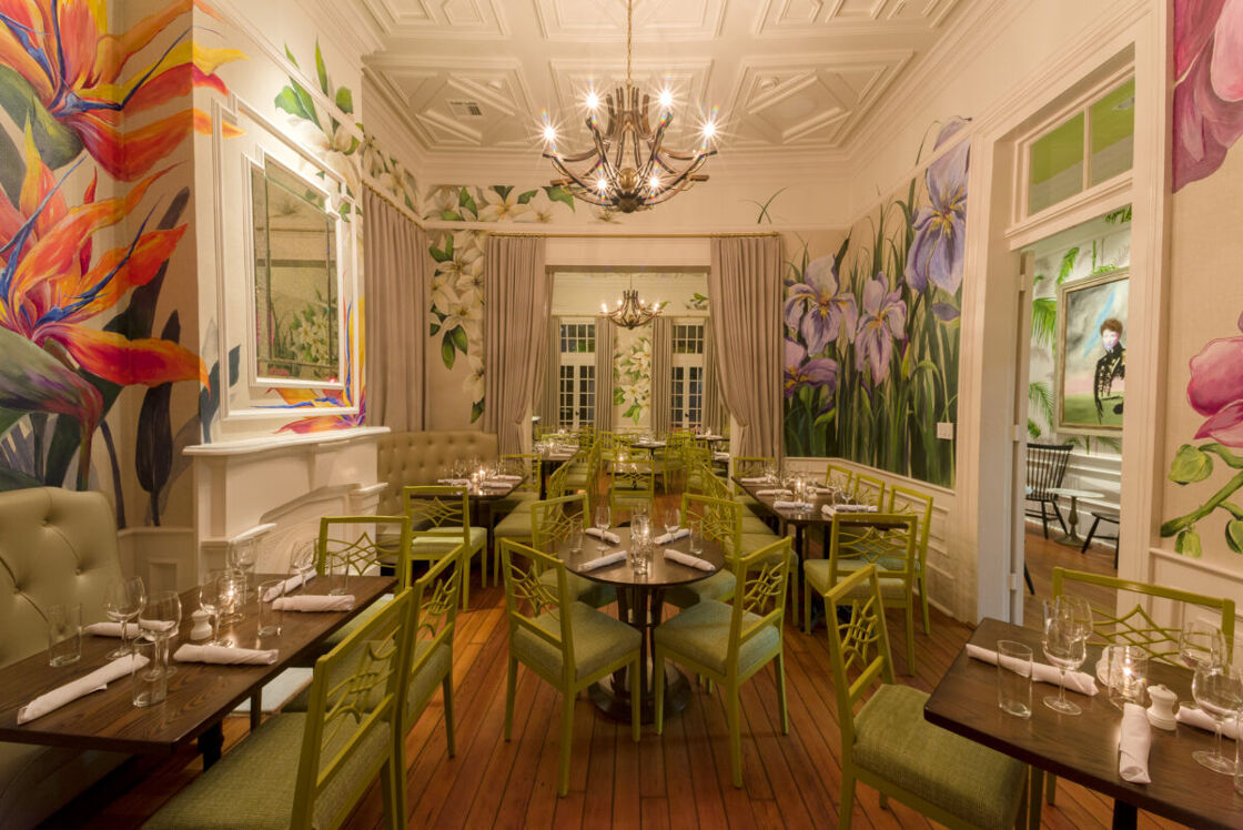 A parlor with several types of flowers painted on the walls. Green chairs sit around several wooden tables set for a meal.