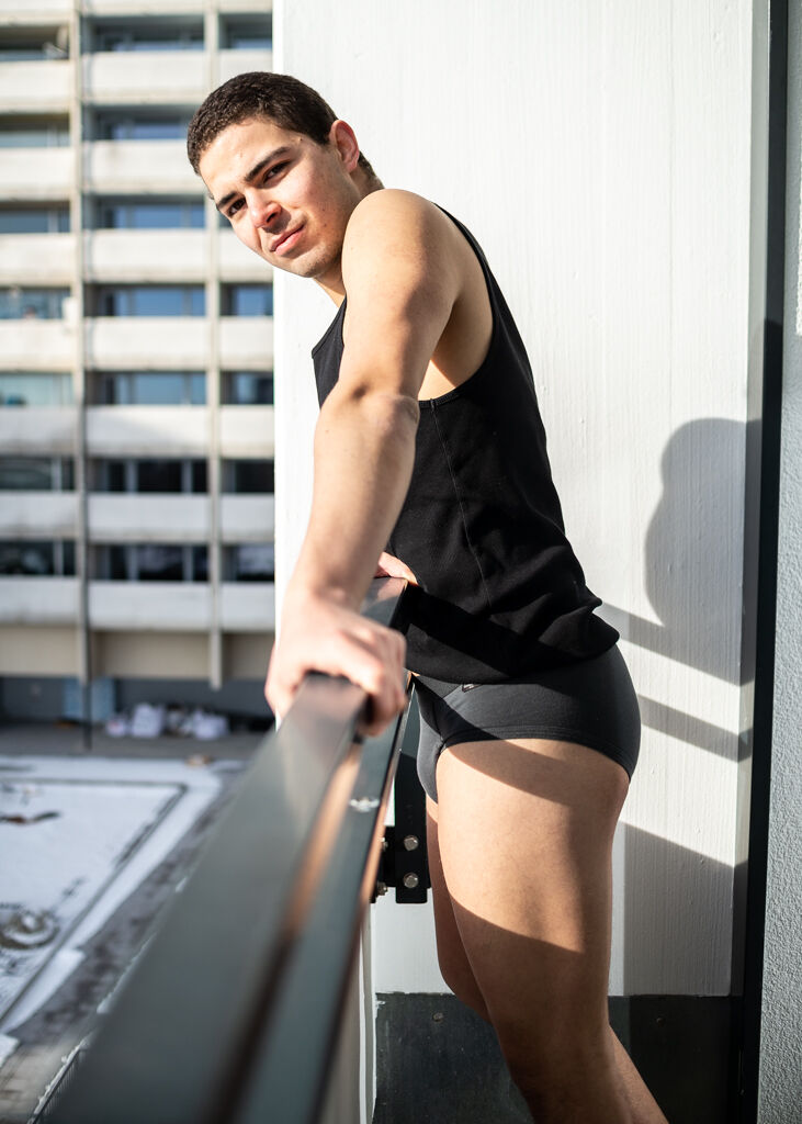 A young man in a black tanktop and black underwear poses on a balcony