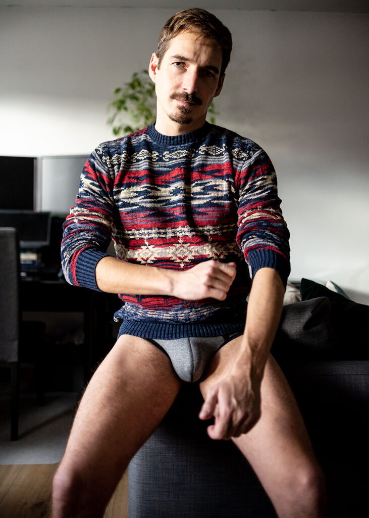 A man with a mustache poses in a blue and red sweater and a jock