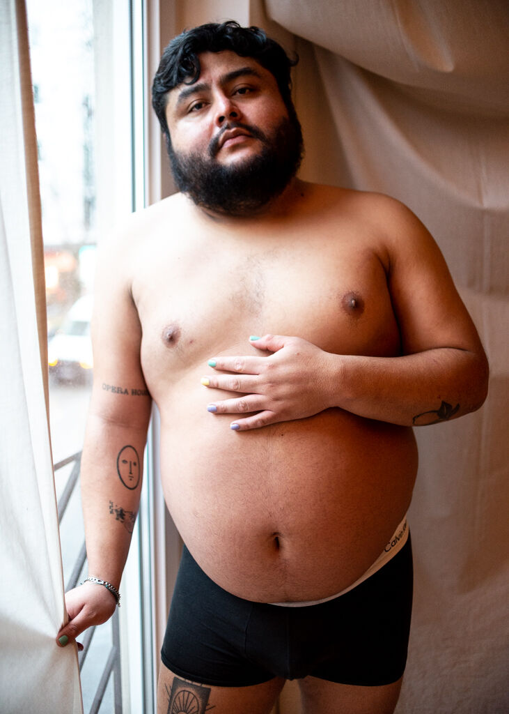 A Latino man with his shirt off and wearing black trunks poses next to a window