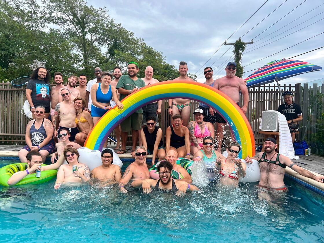 A large group of trans folks pose beside an inflatable rainbow balloon in a pool