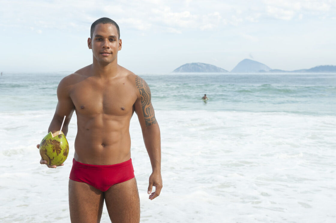 Muscular Brazilian man holding green coconut on the beach in red bathing suit smiling in Rio de Janeiro Brazil