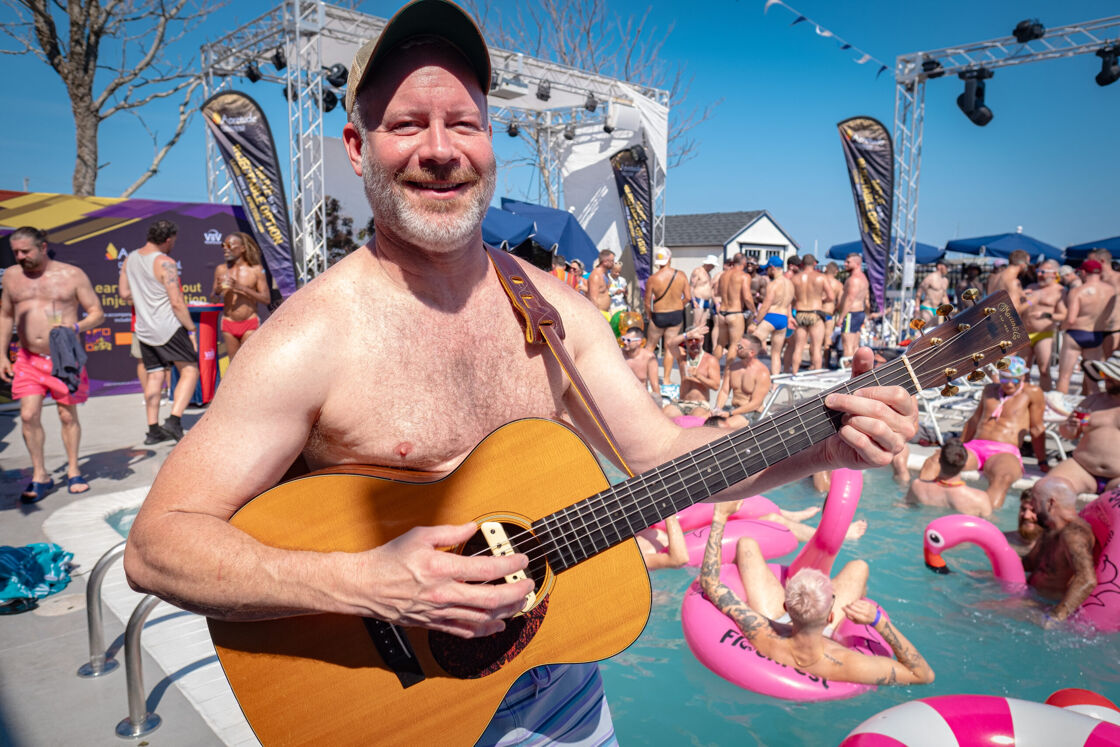 Man playing guitar by the poolside with people in the background.