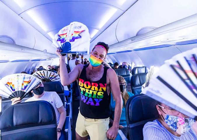 A man stands in the aisle of a plane with a rainbow mask, fan and a shirt that says in rainbow lettering 'born this way'