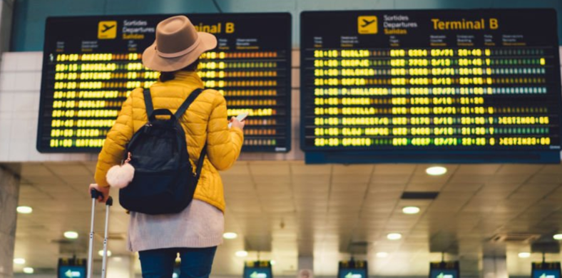 A traveler wearing a black backpack, yellow puffy coat and tan hat stands in front of a departure time board