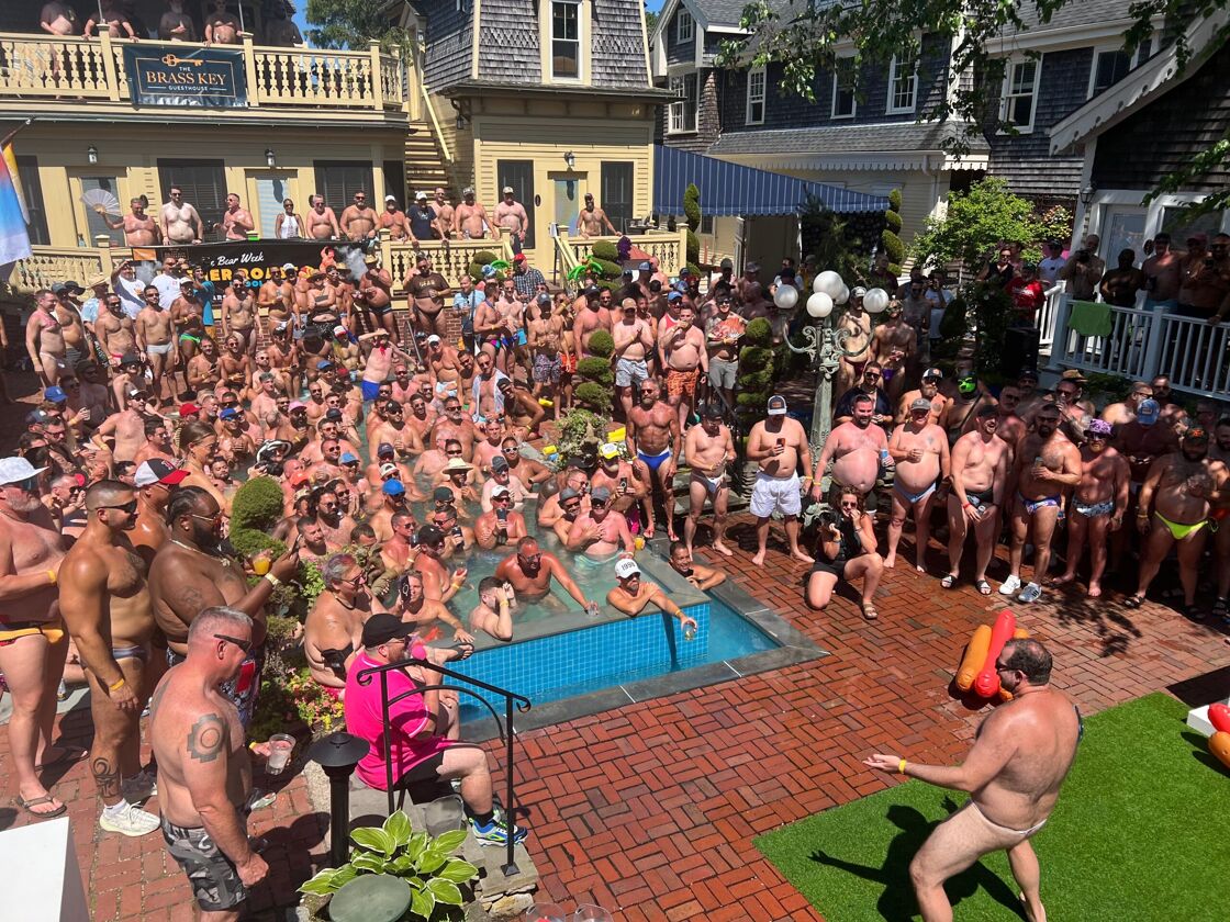 A large group of mostly shirtless men gather at an outdoor pool to watch another man wearing only a jockstrap talk.