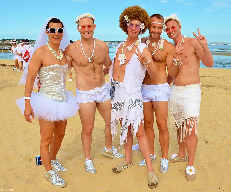 A group of men stand on the beach wearing white accessories and smiling at the camera. Some are throwing up peace signs with their fingers