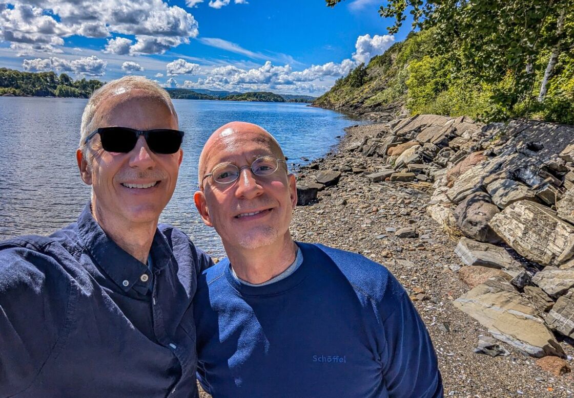 Brent and Michael standing on the beach on a sunny day the fjord behind them. Author and his husband on the island of Hovedøya with the Oslo Fjord behind them