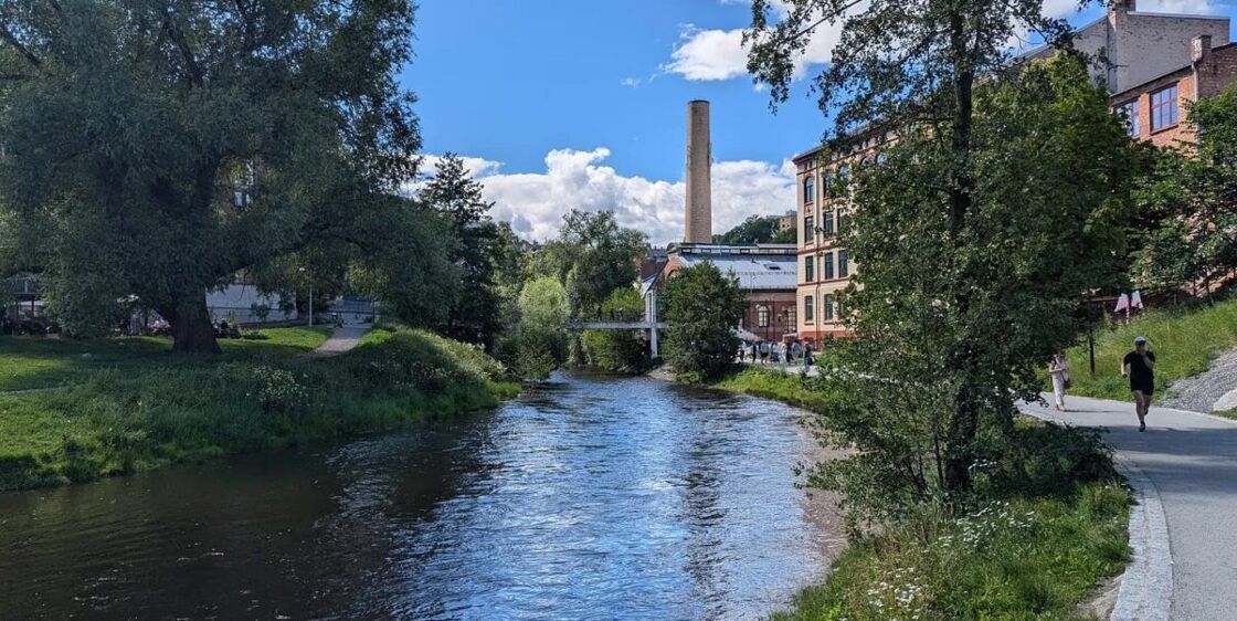 River flowing between tree-lined banks, a walking path on the right, and an old smokestack in the distance. The Akerselva River on a non-rainy day!