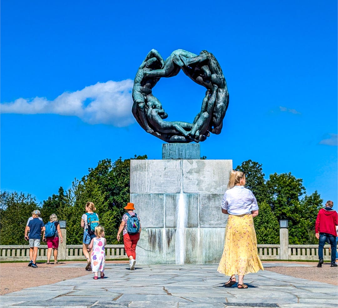 A woman in a yellow dress is right in front of the sculpture, children and other people nearby. A statue at Vigeland Sculpture Park