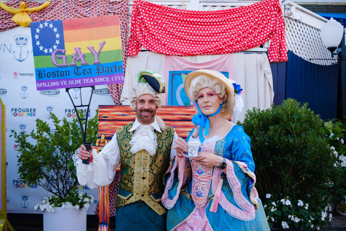 Two people dressed in period garb with colorful makeup stand with a sign that reads "GAY Boston Tea Party"