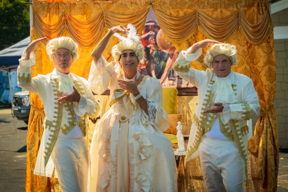 Three men in fine French costumes shimmering in gold smile and pose for the camera