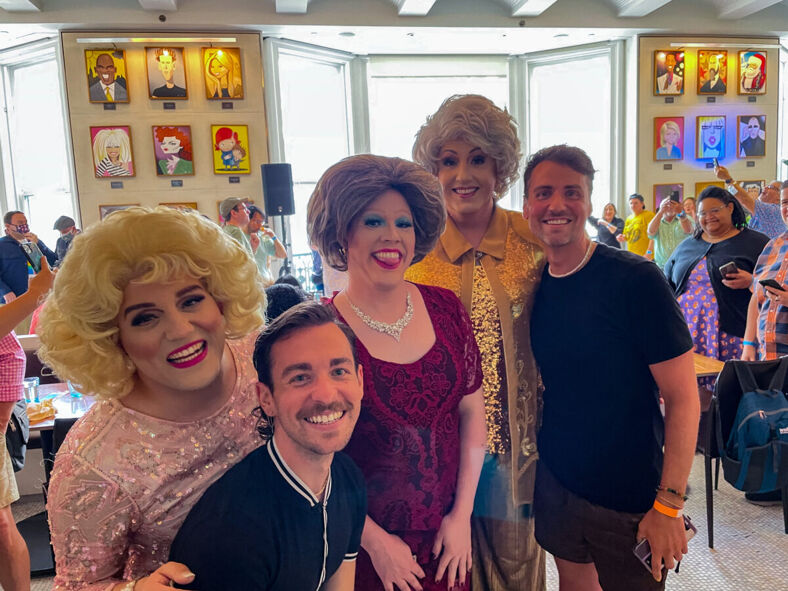 A group photo at a drag brunch featuring three drag performers dressed as the Golden Girls, with two smiling attendees in front of them. The background shows a lively restaurant setting with portraits on the walls.