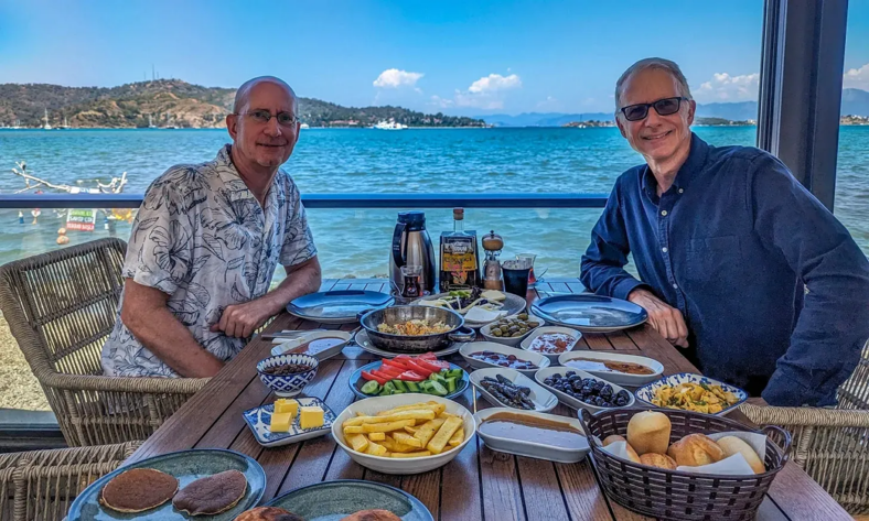 Brent and Michael sit seaside at a table loaded with food