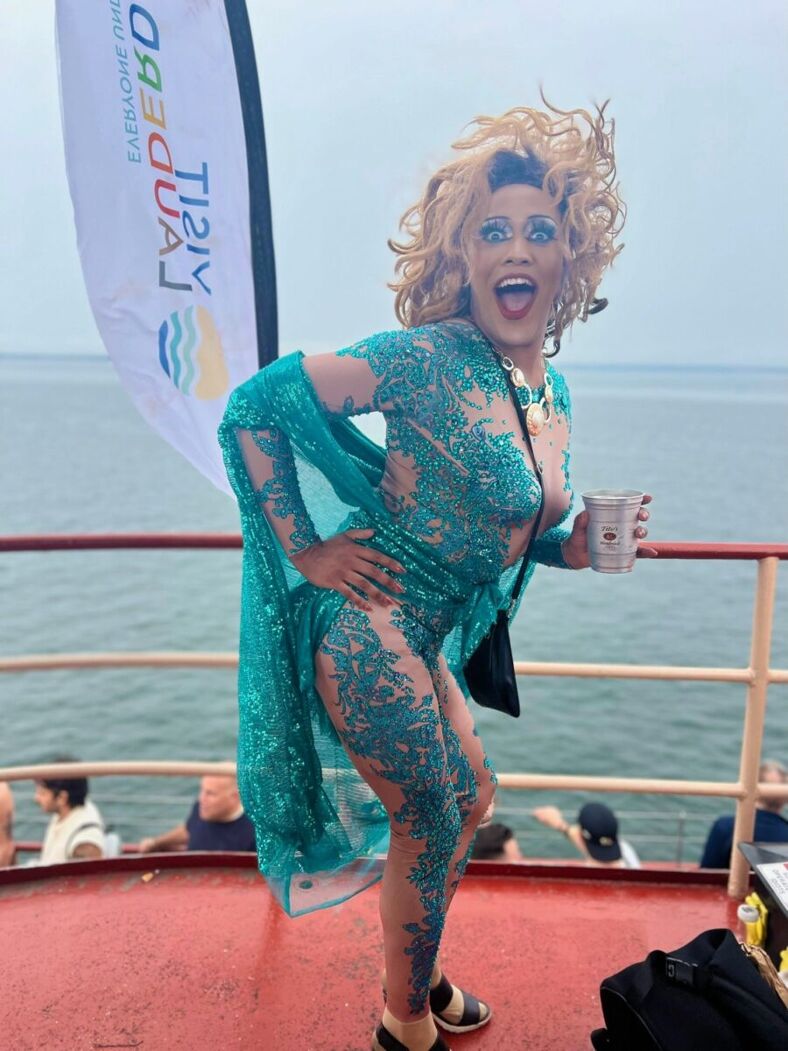 A gorgeous queen in teal sequins poses excitedly for the camera on the dock of a boat