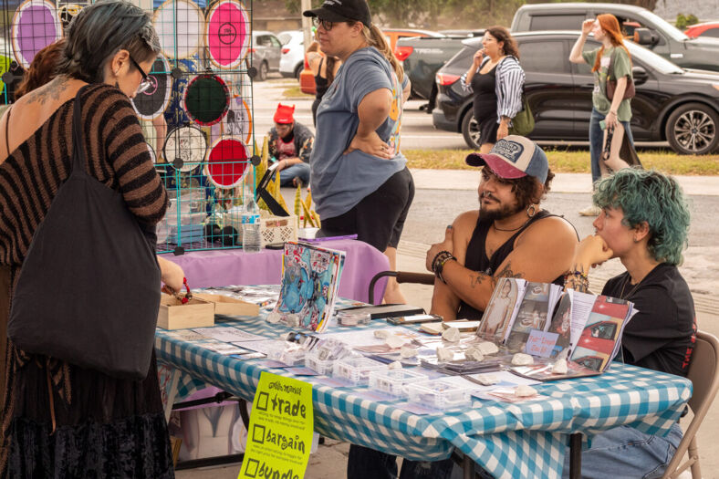 Two people sit at a booth table with their wares in front of them as a few people peruse. 
Photographer: Jordan Macaulay