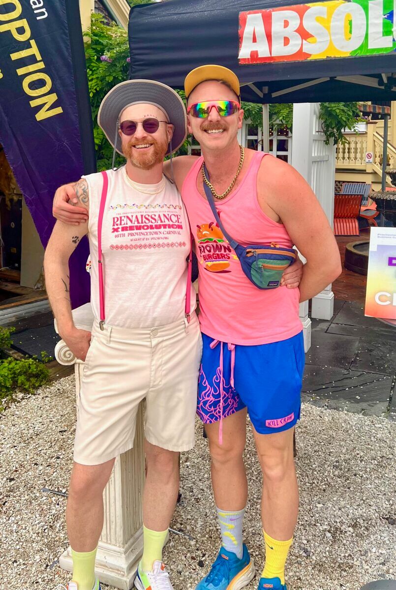 Two men sporting red facial hair and bright clothes smile and embrace for the camera