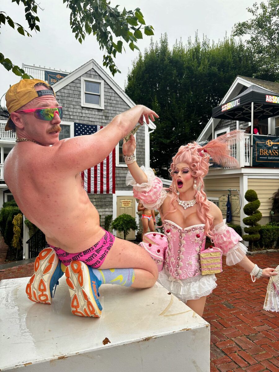 A queen in a bright pink cake couture costume gives a dollar to a shirtless man in a pink Speedo 