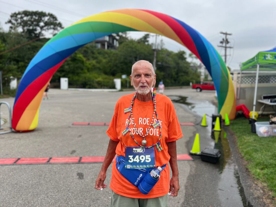 A greying man wears a bright orange shirt that reads 'roe roe roe your vote' and a racing bib pinned beneath. He stands in front of a rainbow balloon arch