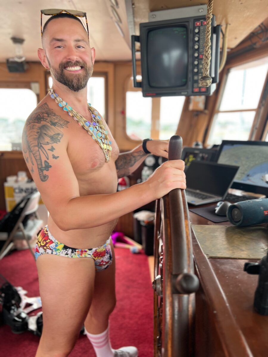A smiling man wearing a colorful speedo and shiny necklace stands at the helm of the ship