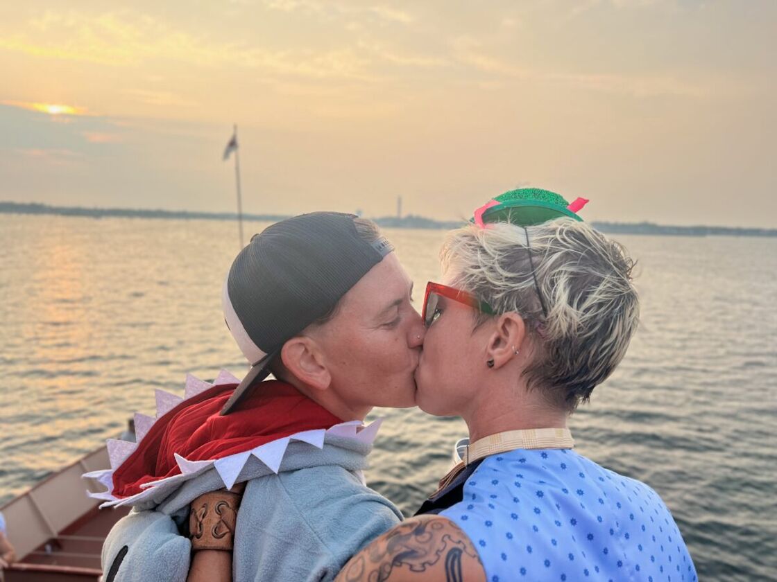 A couple, both with sort hair, one wearing a backwards baseball cap kiss with the water in the background