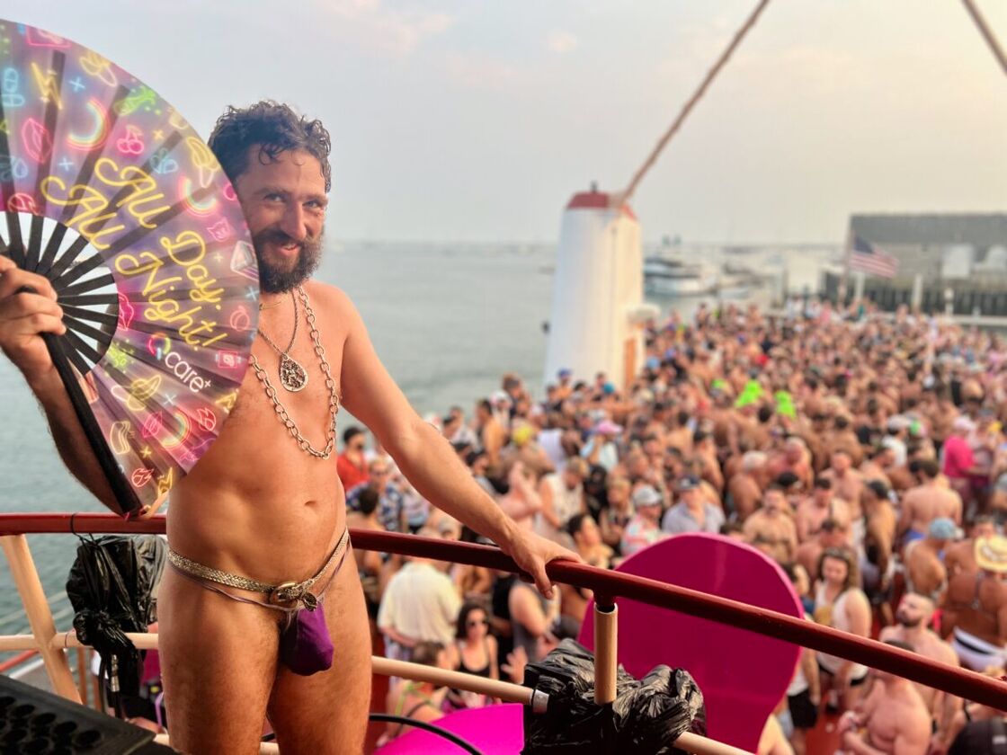 A man stands on a ship wearing only a purple jockstap smiles while holding a bright fan. Below him in the background, the ship is full of people