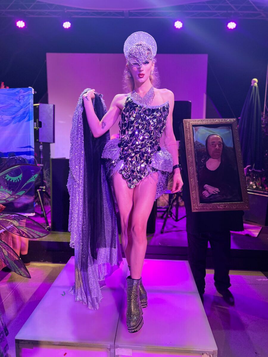 A femme queen walks the catwalk with pieces of art walking behind her