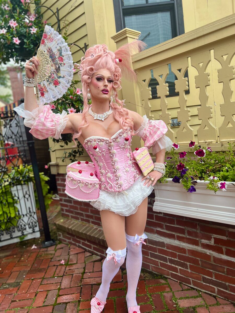 A drag queen dressed in garb riddled with cake and French flourishes strikes a pose with her fan in hand
