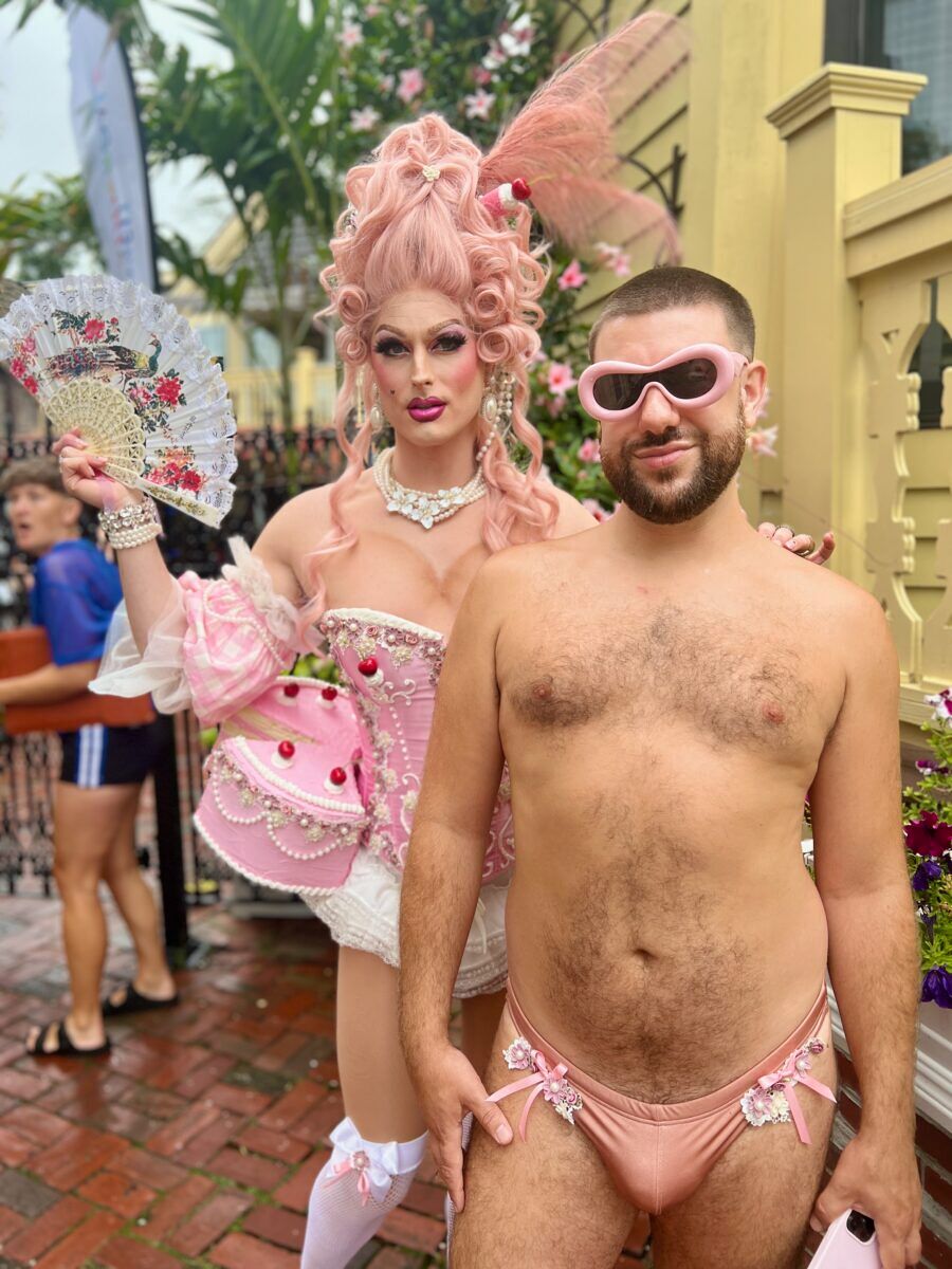Queen of couture in her cake corset stands behind a hairy cutie with a pink jockstrap on